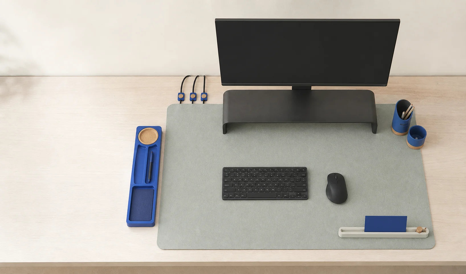 A measured home-office desk arranged with a desk mat, low platform, trays, cable guides, and paper references in Fieldlay's bone, graphite, mist, and cobalt palette